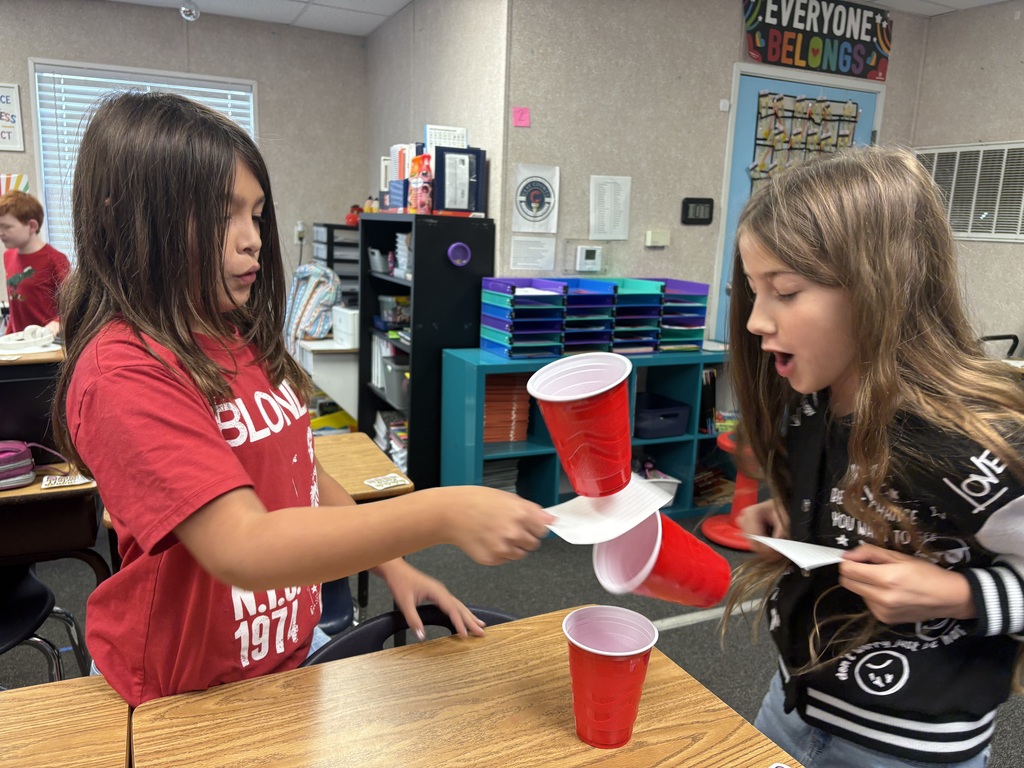 Two girls in a classroom setting. One holds cups, the other attempts to stack them.