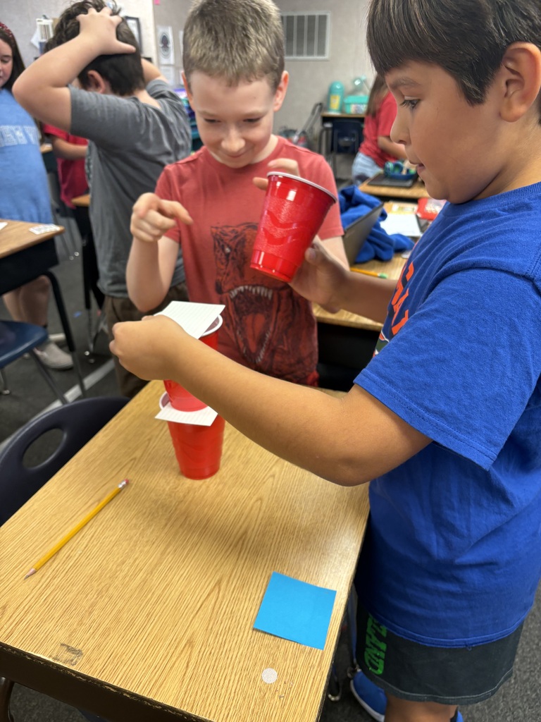 Two young boys are working together at a table, holding red cups, and doing an experiment.