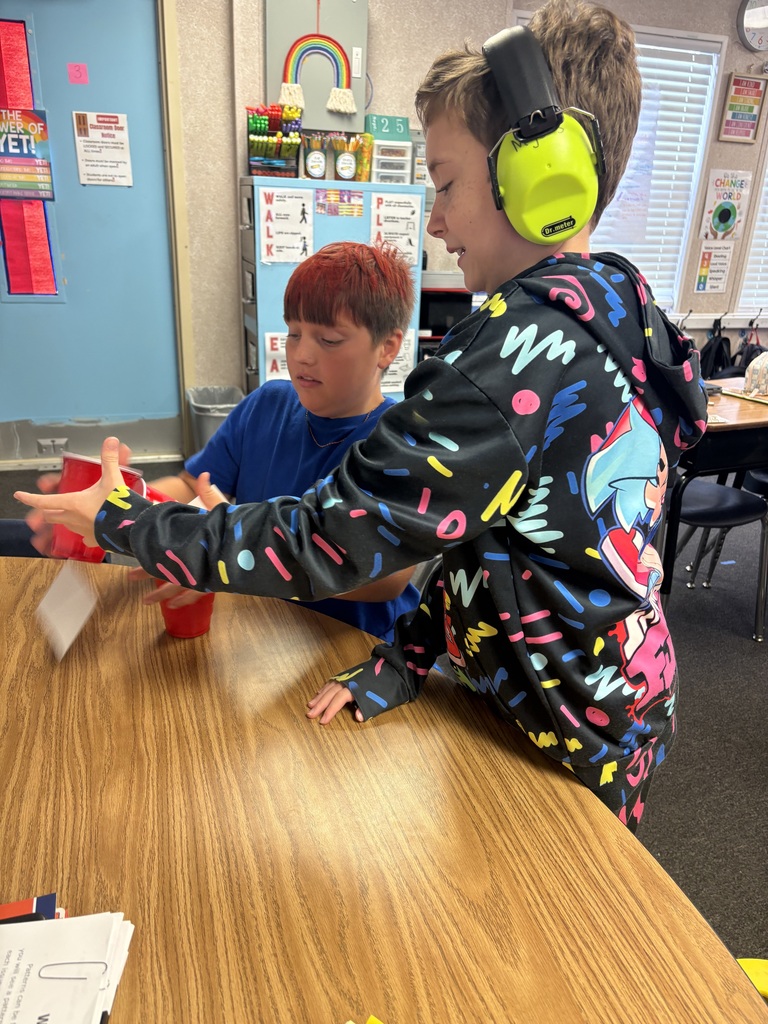 Two boys, one with headphones, sit at a table, sharing a red cup and papers. Behind them is a room with tables, chairs, and a bulletin board.