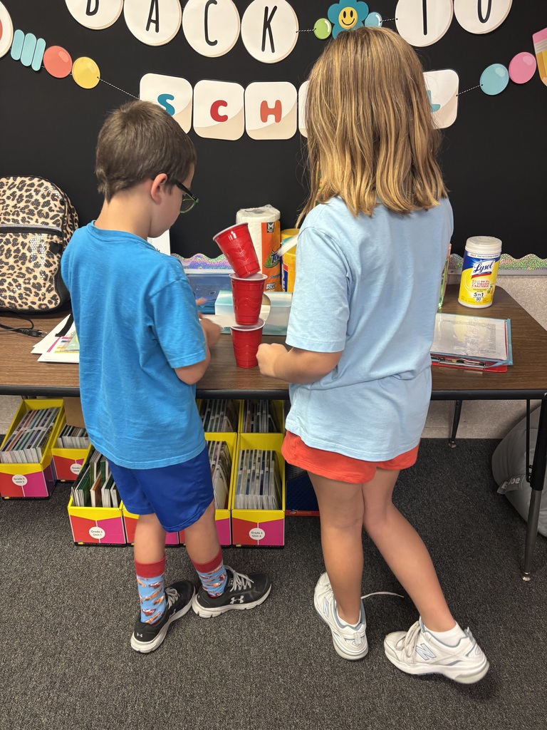 Two children stand by a table, one arranging cups, the other observing. They wear light blue shirts and shorts. A colorful banner adorns the background.