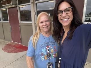 Two women standing outside a building with glass doors, one in a blue shirt with a design and the other in a navy blue dress.