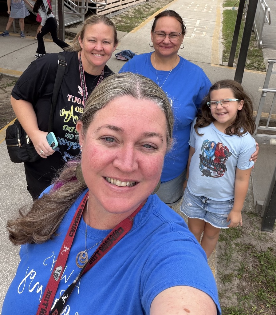 Four people stand on a sidewalk. A woman in a blue shirt smiles at the camera. A young girl, wearing glasses, stands next to her.