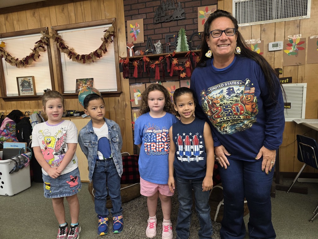 A woman stands with four children in a classroom with wooden walls and decorations. One child wears a denim jacket.