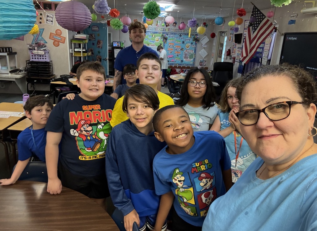 Classroom with students standing behind a table, smiling at the camera. One adult stands in front of them, wearing glasses and a blue top.