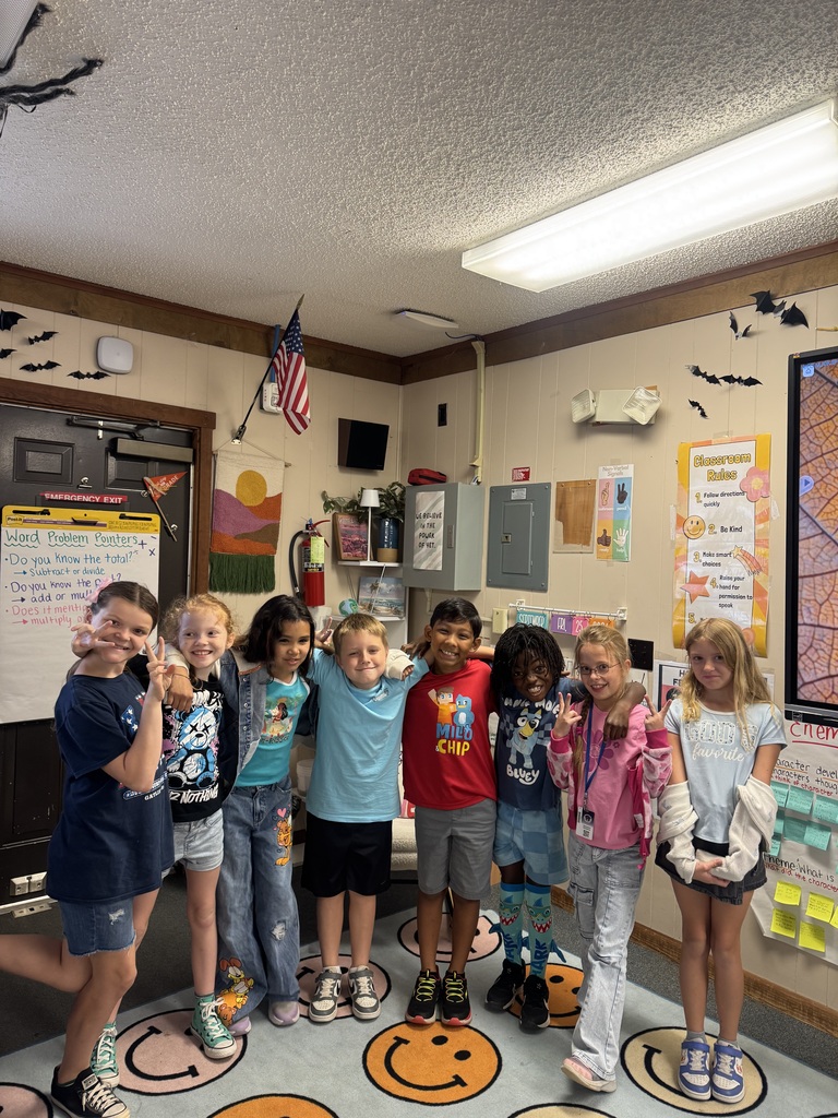Several children in a classroom are standing in a circle, smiling. Some are making gestures.