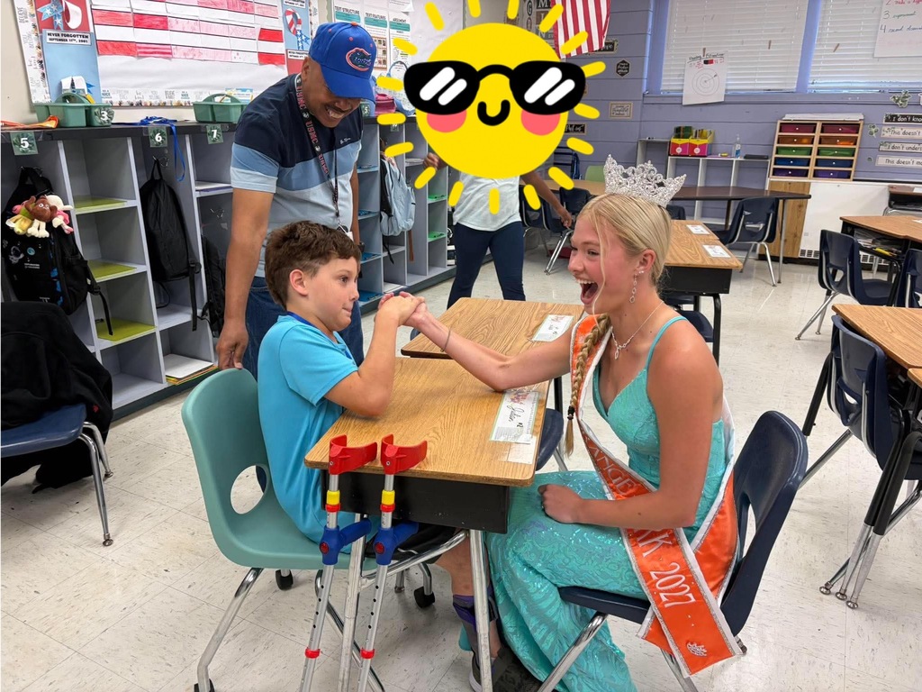 A classroom with tables and chairs; a woman in a sash smiles while a boy holds her hand.