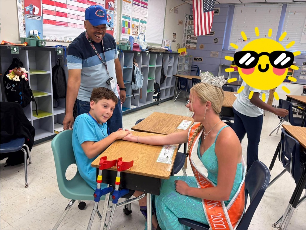 A woman and a boy sit at a desk in a classroom, shaking hands. A woman wears a sash.