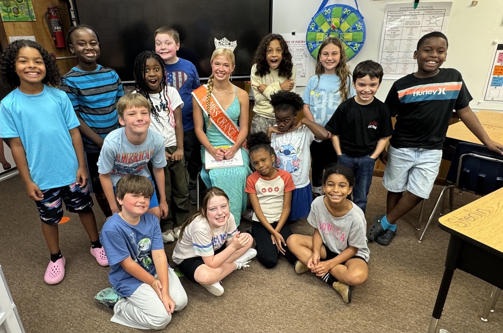 Kids in a classroom, one wearing a sash, posing for a group photo with a teacher.