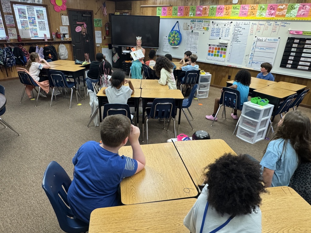 A classroom with young students sitting at tables facing the front, where a teacher is present.