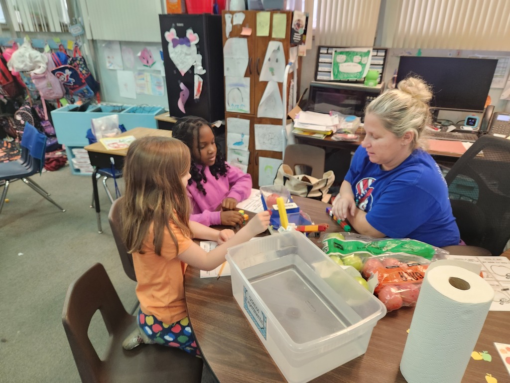 Two children, one in pink and one in orange, work at a table with a woman in blue.