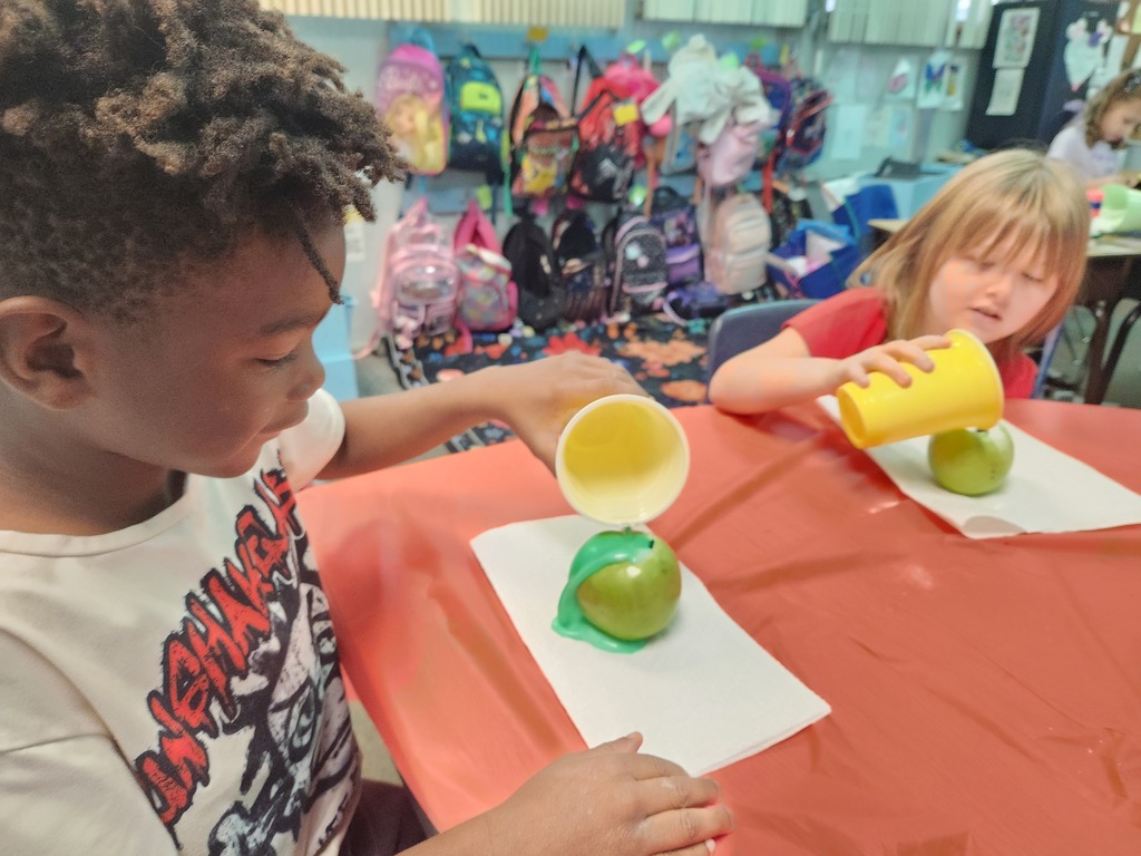 Two children use cups to pour liquid over apples on paper plates at a table.