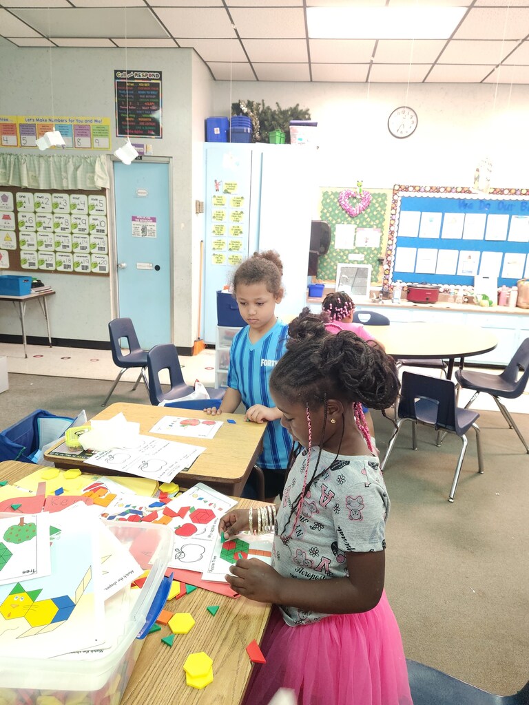 Two young children, one in a pink skirt, work together at a desk in a classroom.