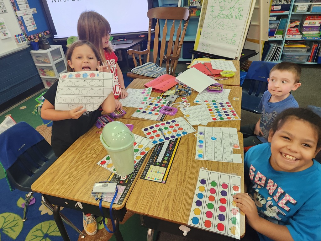 Four children sit at desks, smiling, with one holding a paper and another a plastic container. Colorful materials and papers are on the table.