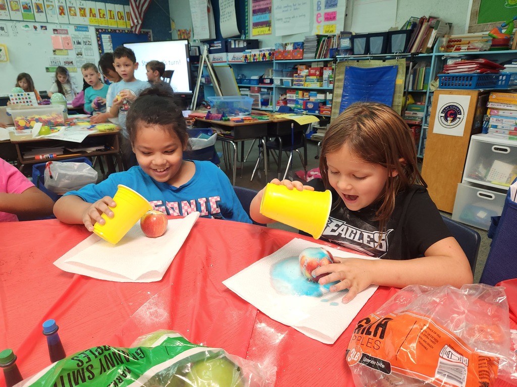 Two young girls seated at a table, engaging in an activity with yellow cups and an apple.