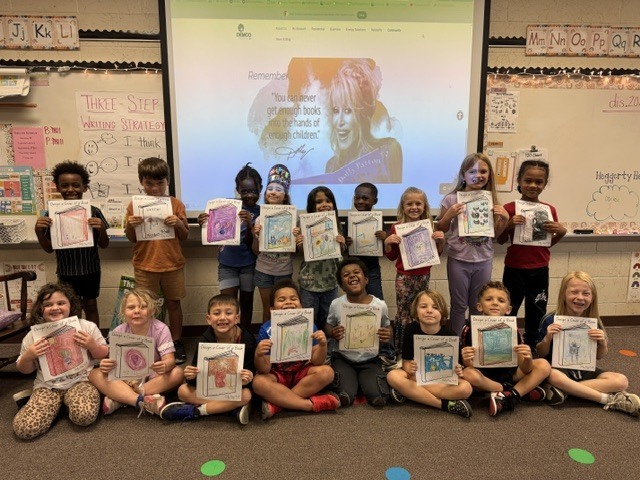 Students in a classroom pose with drawings, a projector screen displays a woman in the background.