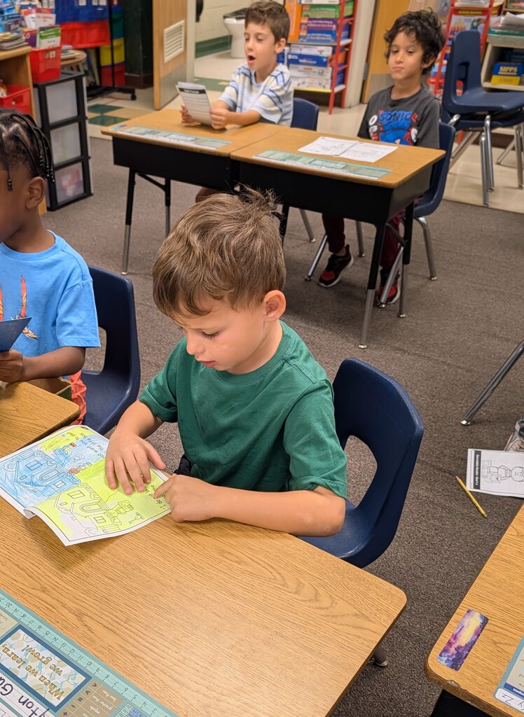 Four children in a classroom. Three sit at desks with reading material, one looks at a tablet.