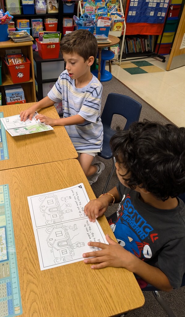 Two children seated at a table, engaged in coloring activity. One holds a paper with a house, the other holds a paper with a rocket.