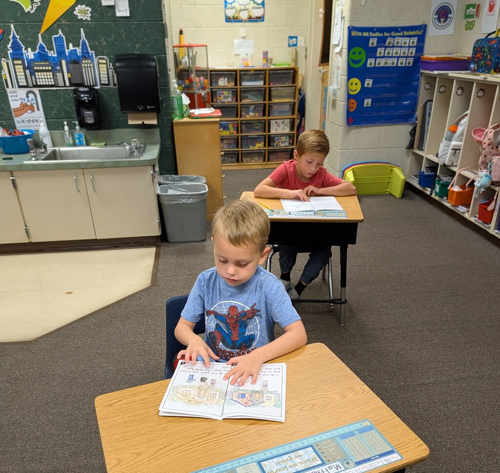 Two children in a classroom, one reading a book while another sits at a desk. Behind them are shelves and a sink.