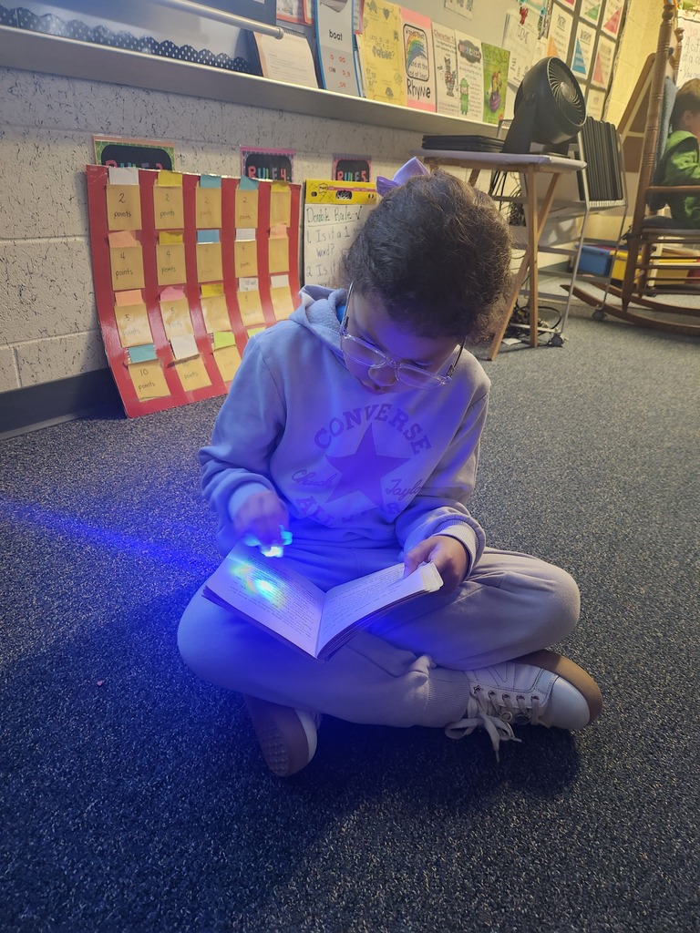A child in glasses sits on the floor, reading a book with a purple flashlight.