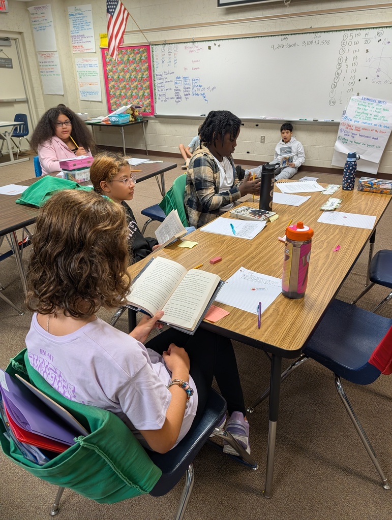 Students seated at tables in a classroom, engaging in reading and writing activities. A whiteboard is in the background.