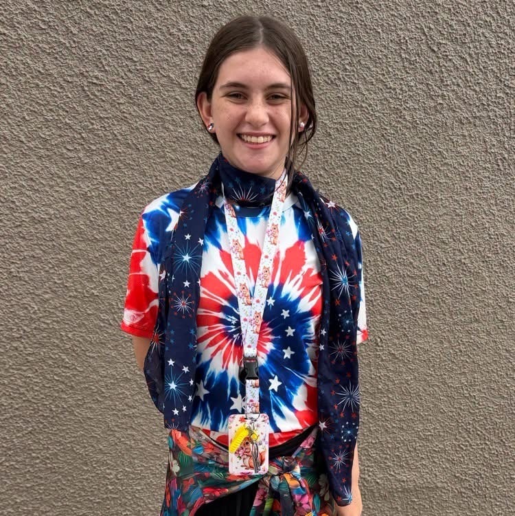 A person stands against a plain wall wearing a tie-dye t-shirt and a scarf with an American flag design.