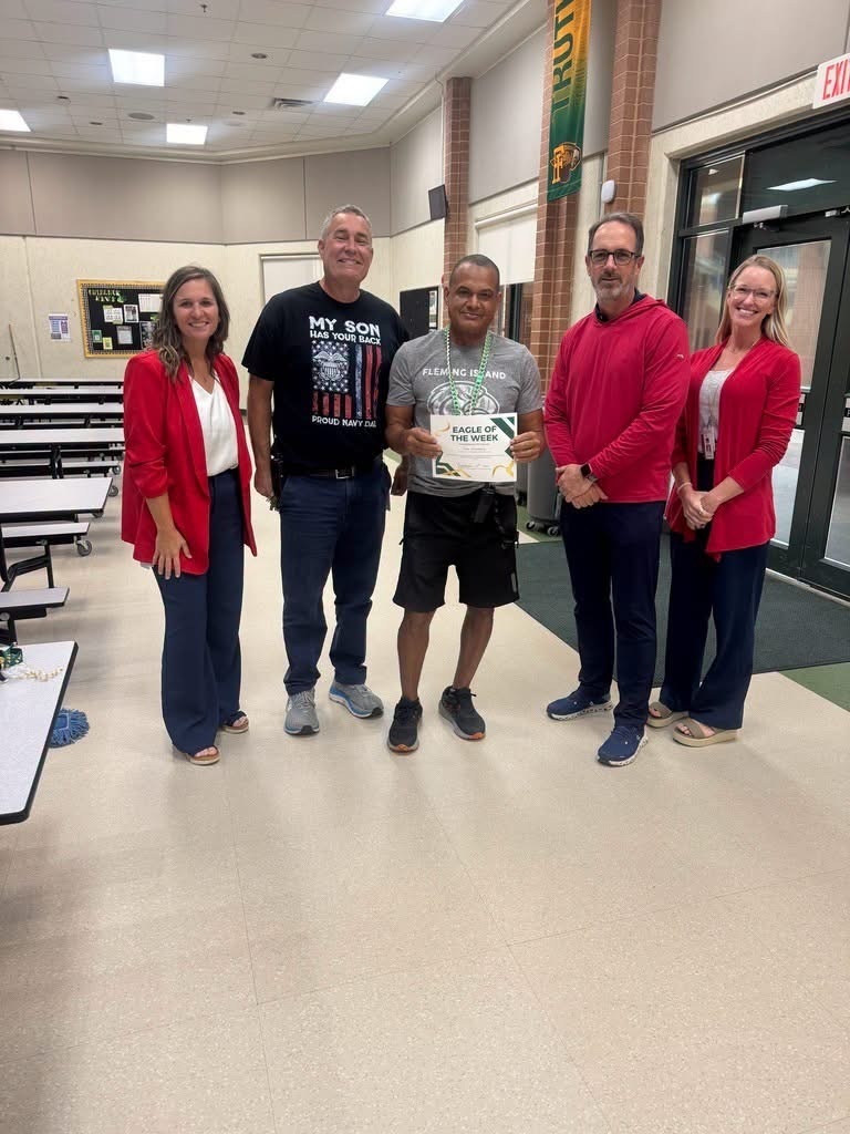 Five adults in a school cafeteria, one holding a framed award, stand with tables and chairs in the background.