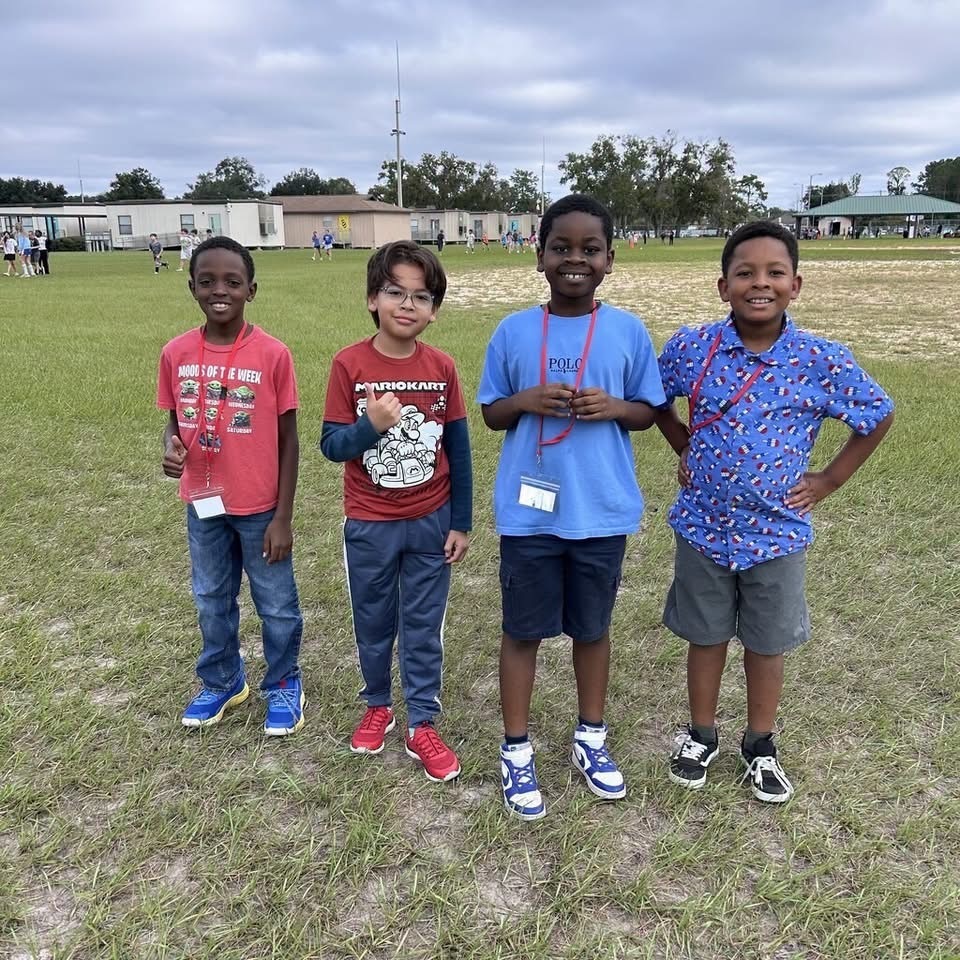Four children stand together on a grassy field. One wears a red shirt, another blue. They all have name tags.