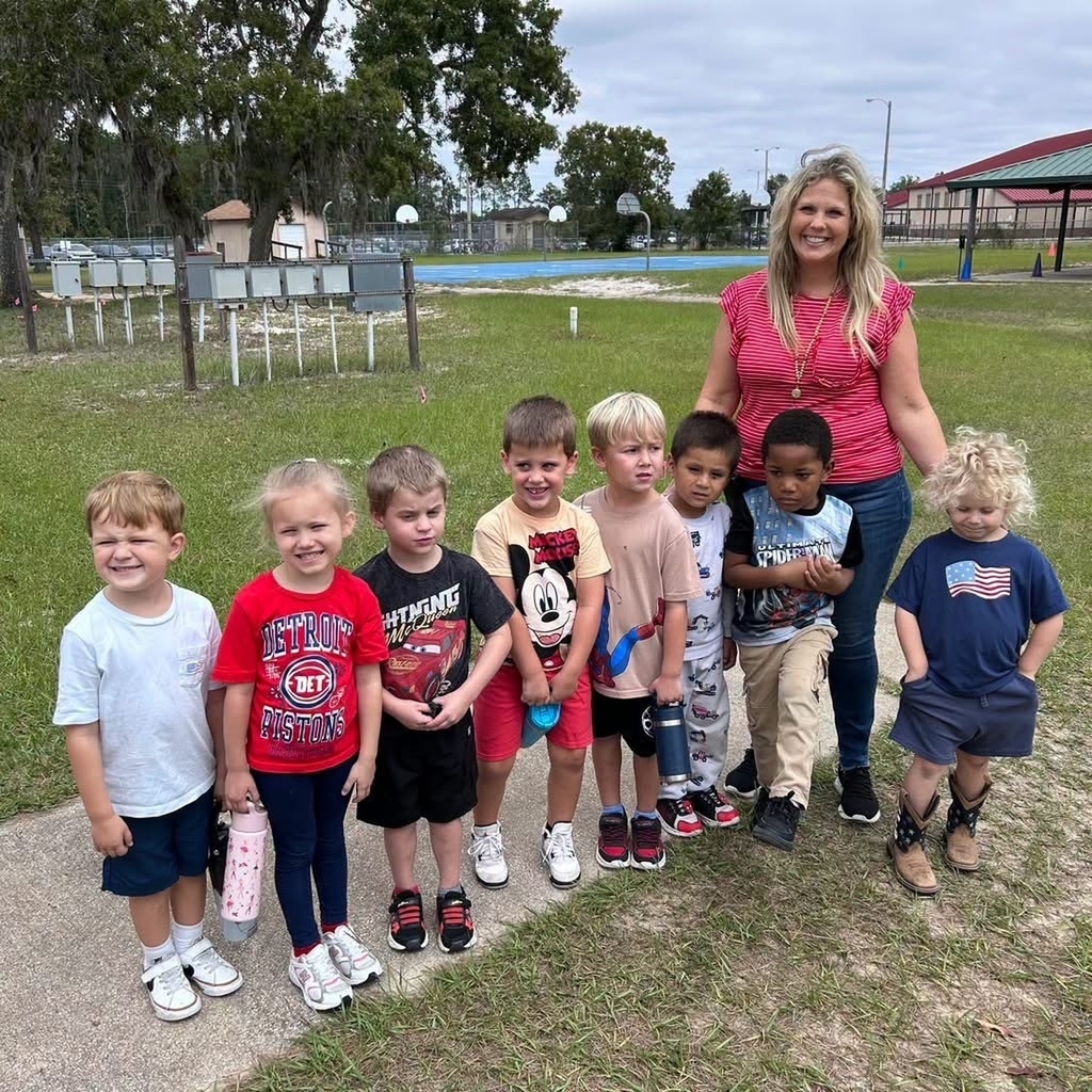 A woman stands with a group of young children in a park area, with grass, trees, and a shelter.