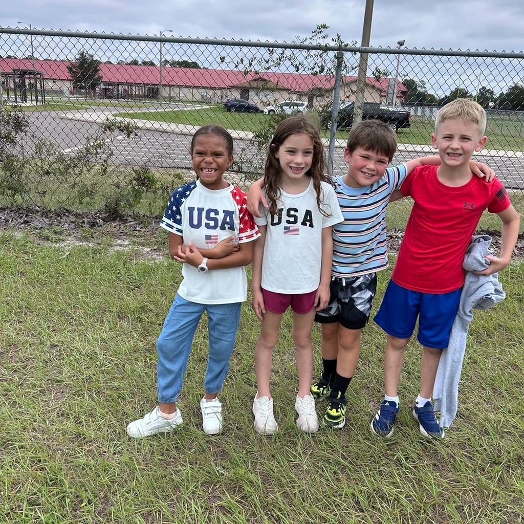 Four children in casual clothes pose in a grassy area behind a fence. Behind them are buildings and a road.