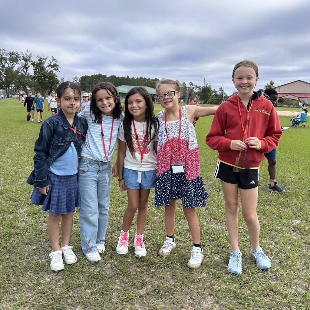 Five girls in casual attire stand on a grassy field. They wear sneakers and one has a lanyard.