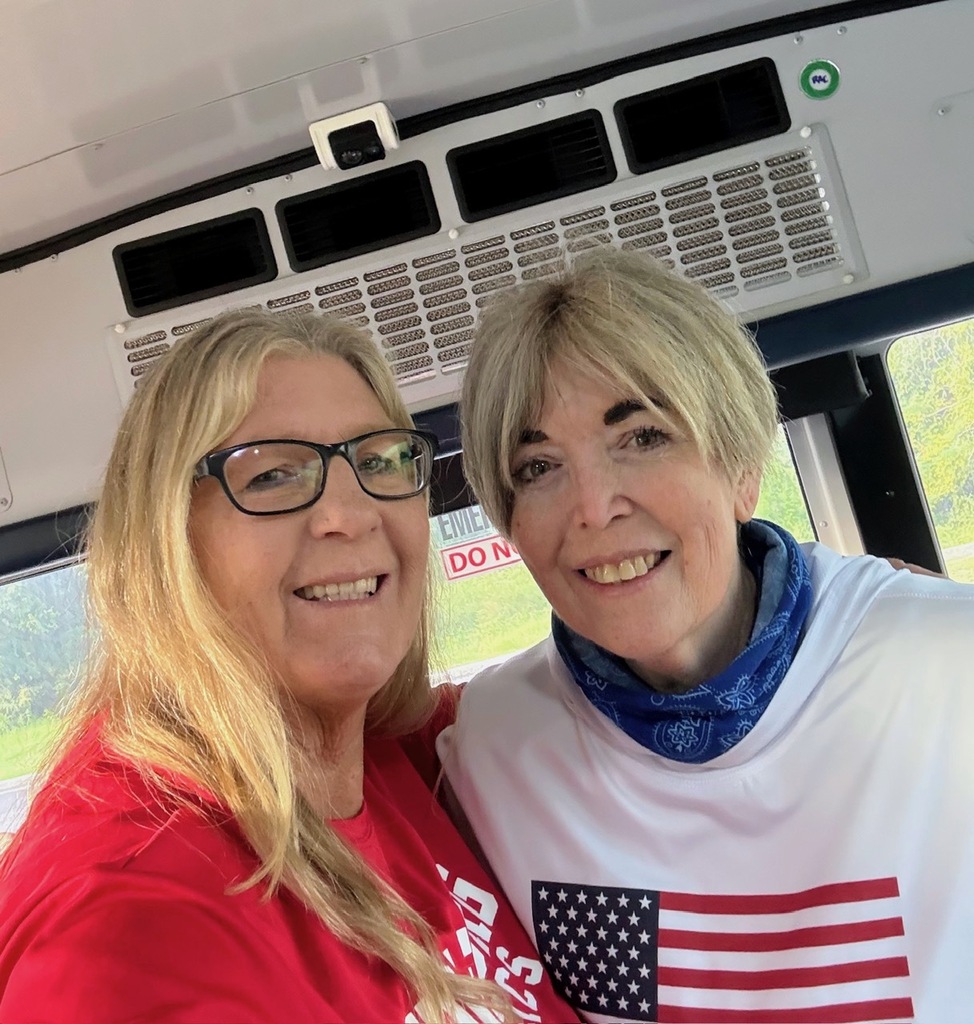 Two women in a bus, one wearing glasses and a red shirt, the other in a white shirt with an American flag.