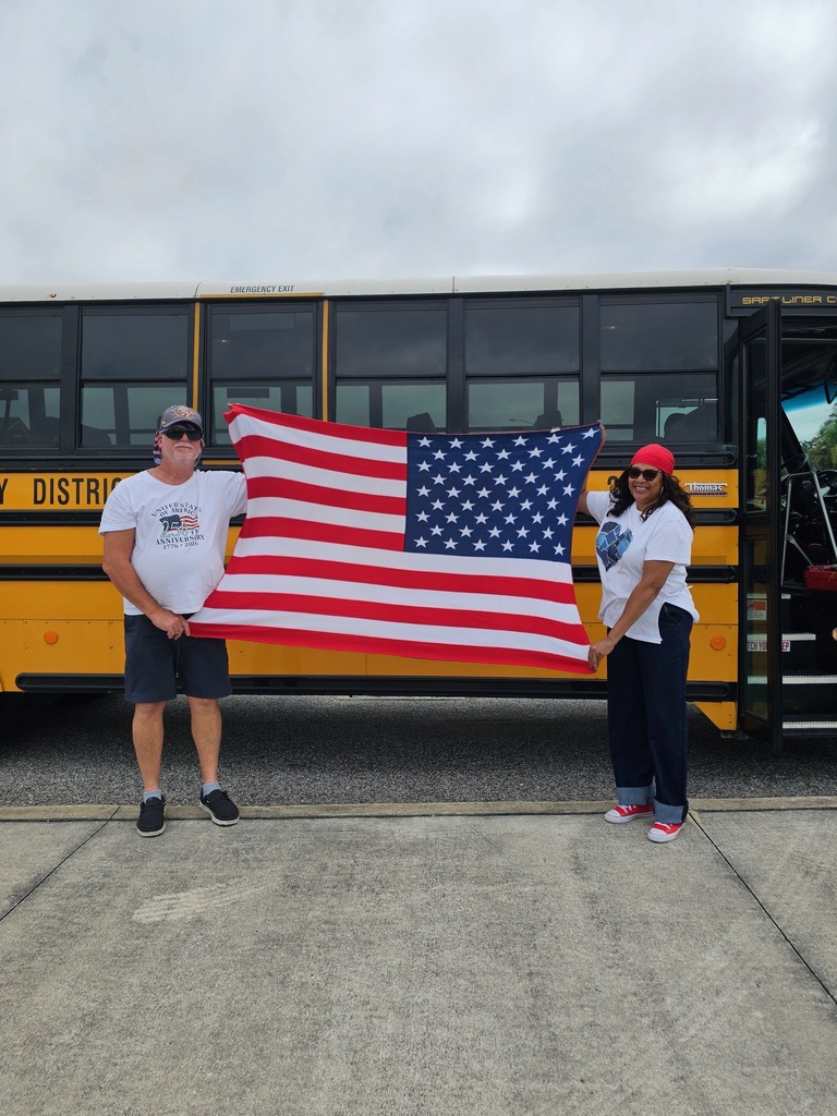 Two people hold a large US flag in front of a yellow school bus.