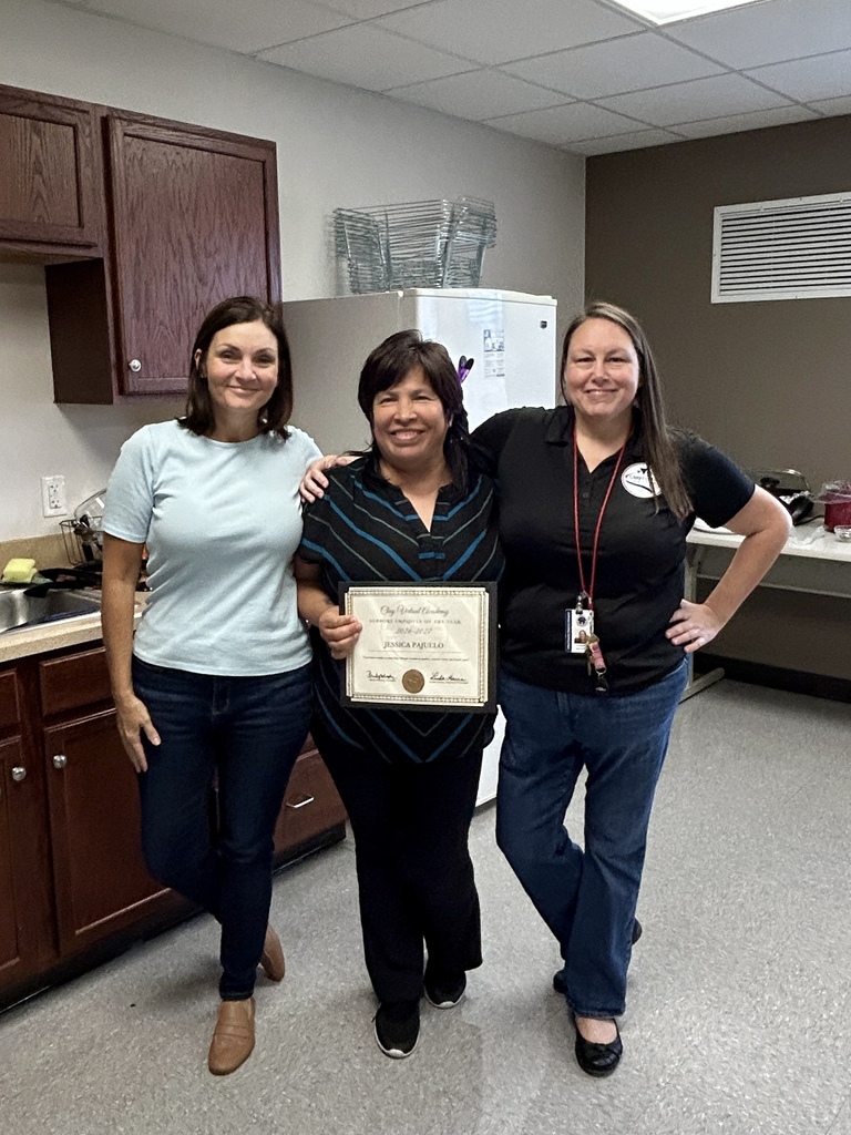 Three women in jeans and shirts stand in a kitchen. One holds a framed certificate. Behind them, a refrigerator and wooden cabinets are visible.