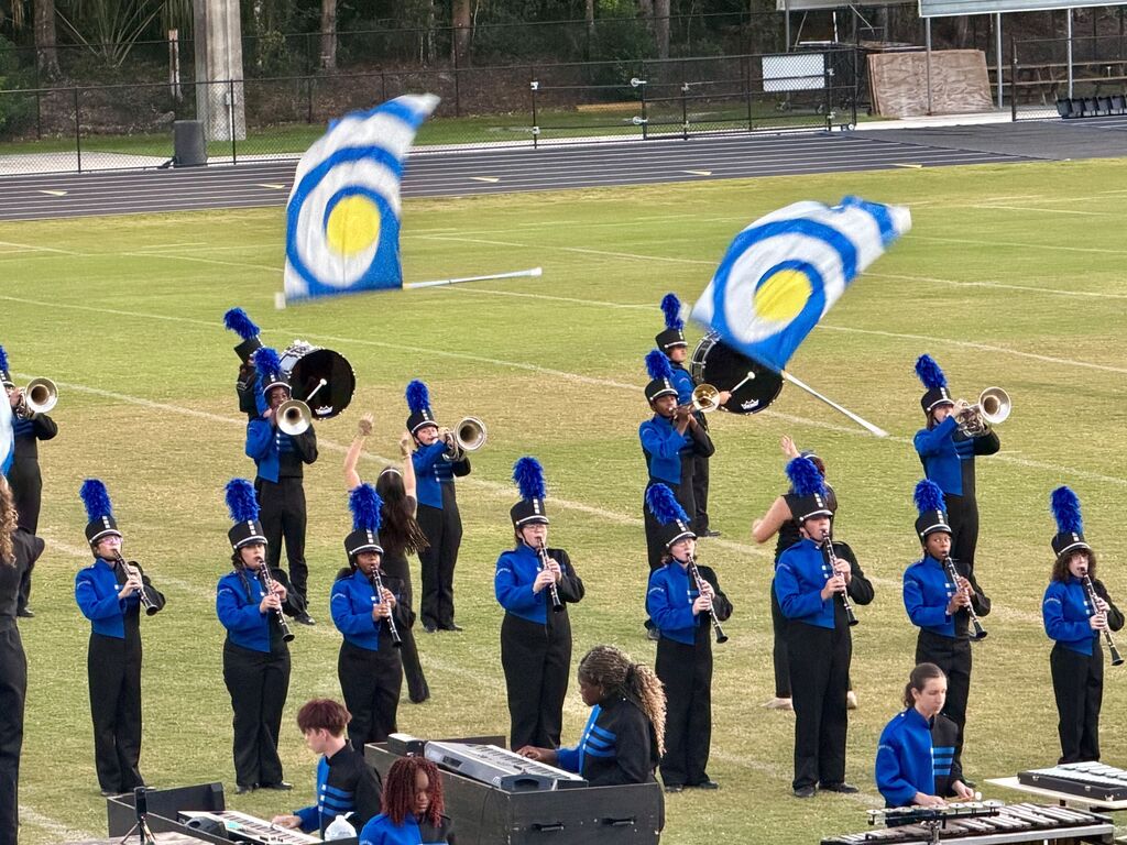 A marching band on a field, with players in blue uniforms, holding instruments. Two flag bearers hold flags with circular designs.