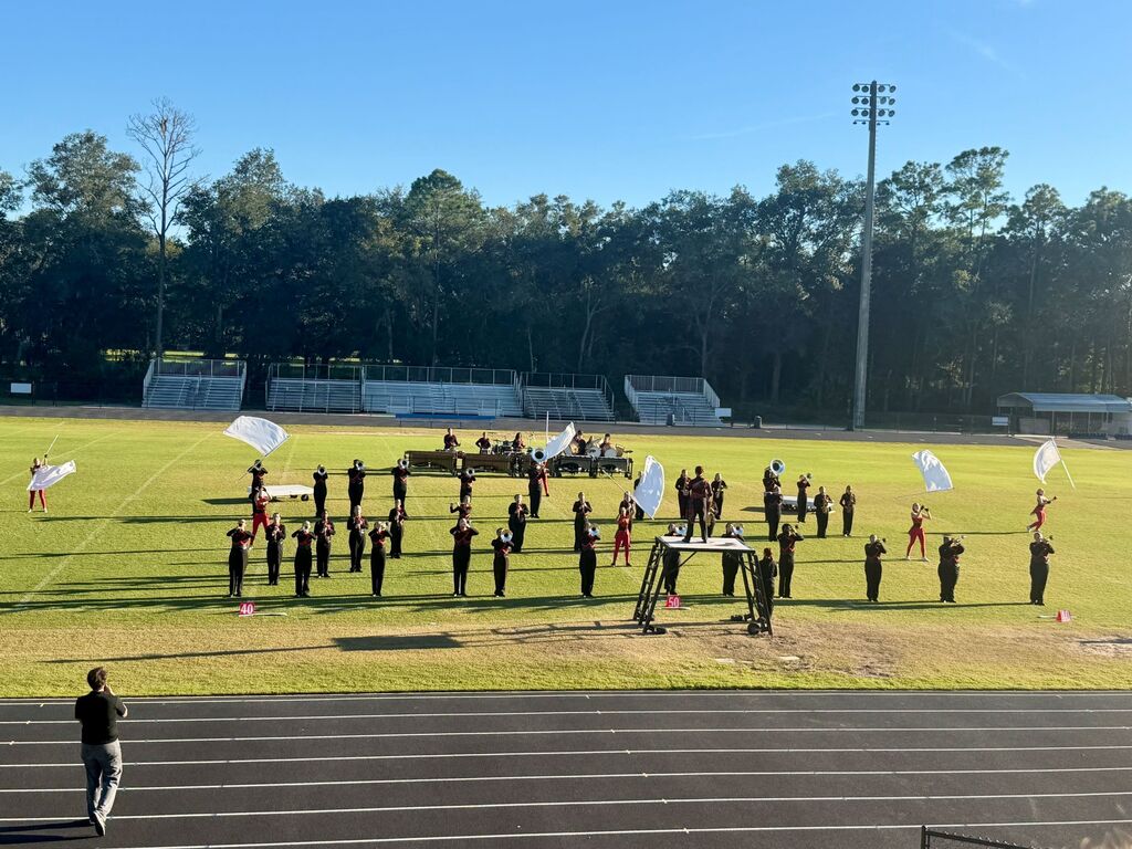 A marching band on a track, holding flags. A person walks near the track. Trees and bleachers are in the background.