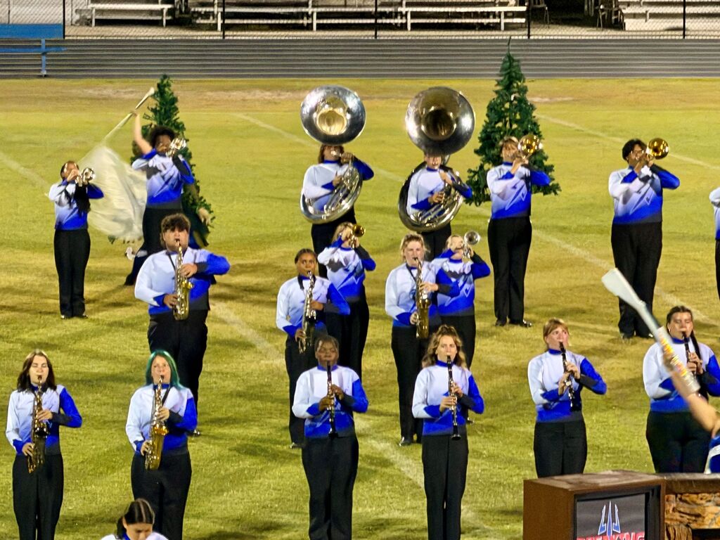 A marching band performs on a field with musical instruments. Members wear blue and white uniforms. Background features trees and bleachers.