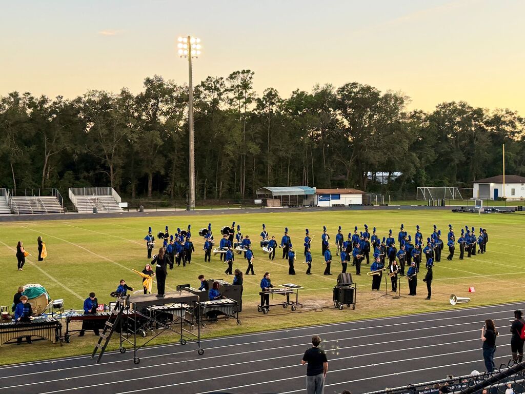 A group of individuals in blue uniforms gathers on a field, with musical instruments and bleachers. Trees surround the open field.