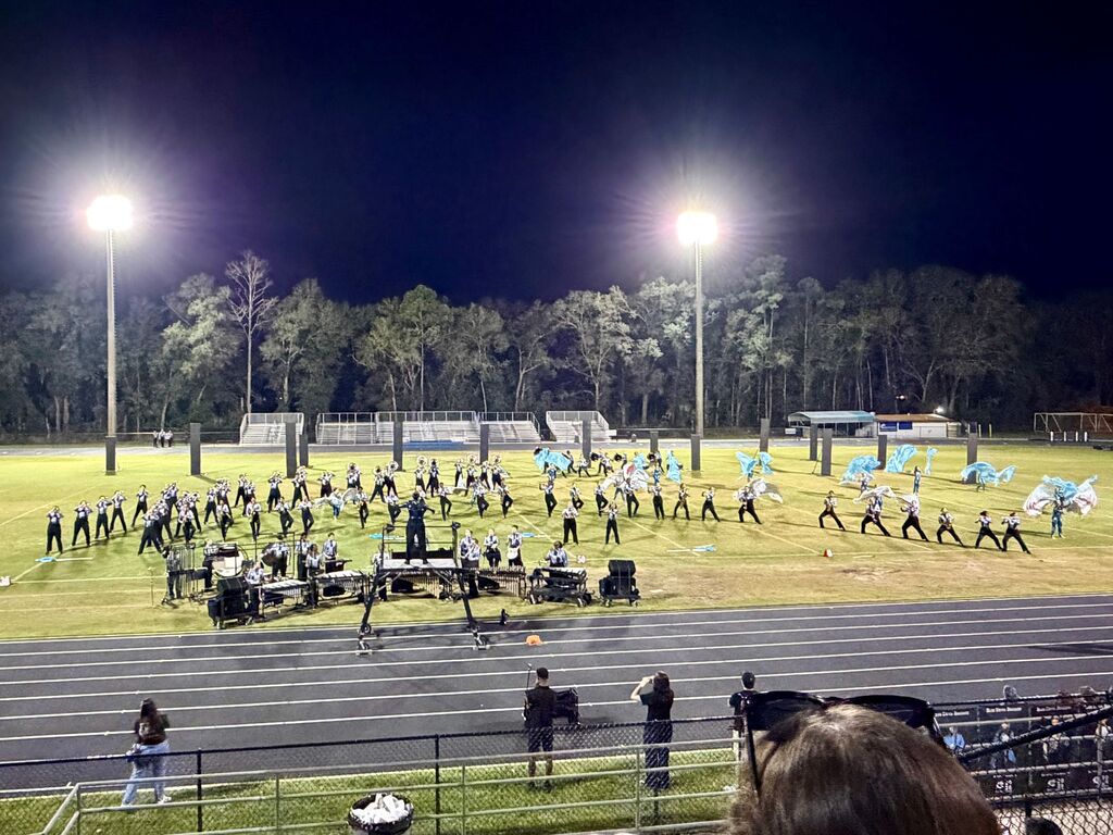 A marching band practices on a field at night. The field is lit by bright lights. Trees are in the background.
