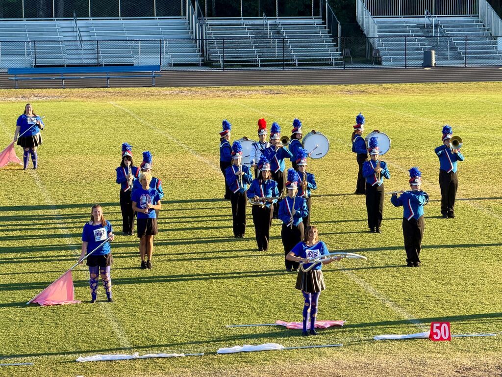 A marching band stands on a field, wearing blue uniforms and hats, playing various instruments and holding flags.