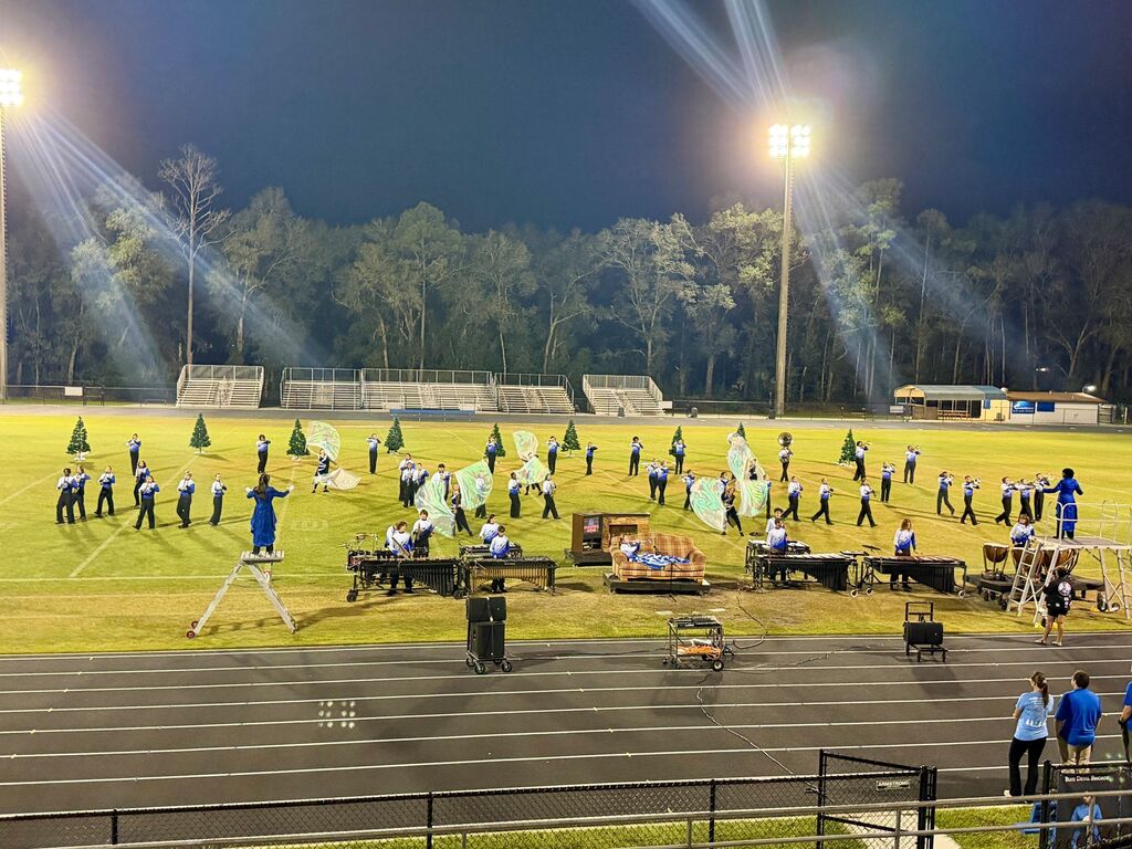 An outdoor field with people performing. A marching band with instruments on a track. Trees and lights in the background.