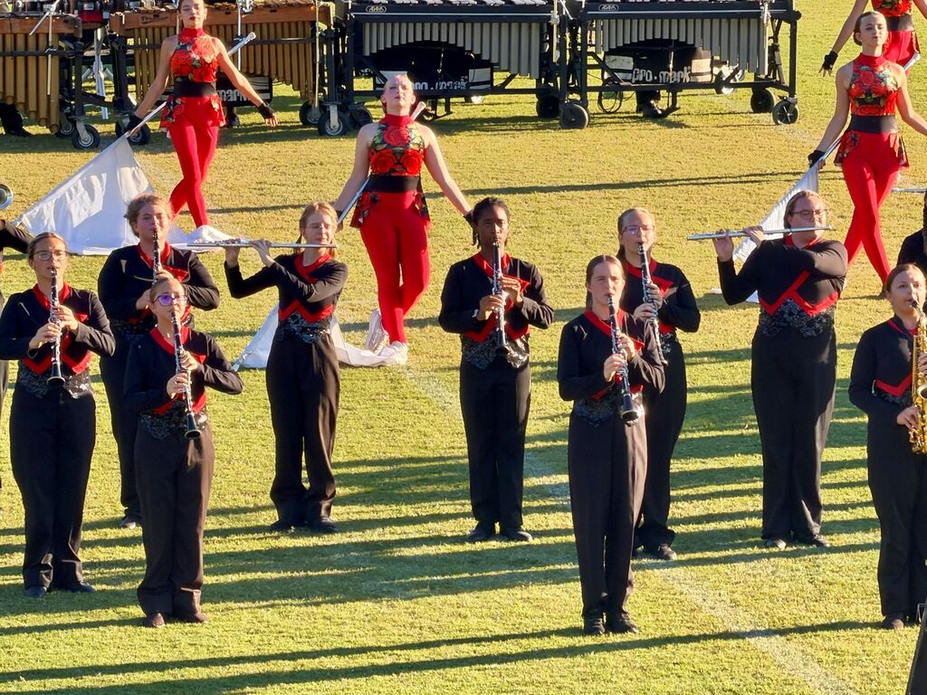 Musicians in black and red uniforms perform on a grassy field, holding instruments. A few players wear white shoes.