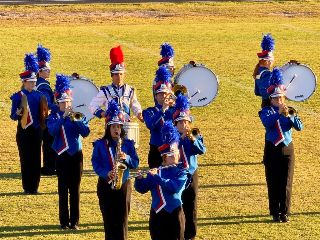 A marching band performs outdoors. Musicians play trumpets, saxophones, and drums. They wear blue and black uniforms with matching hats.