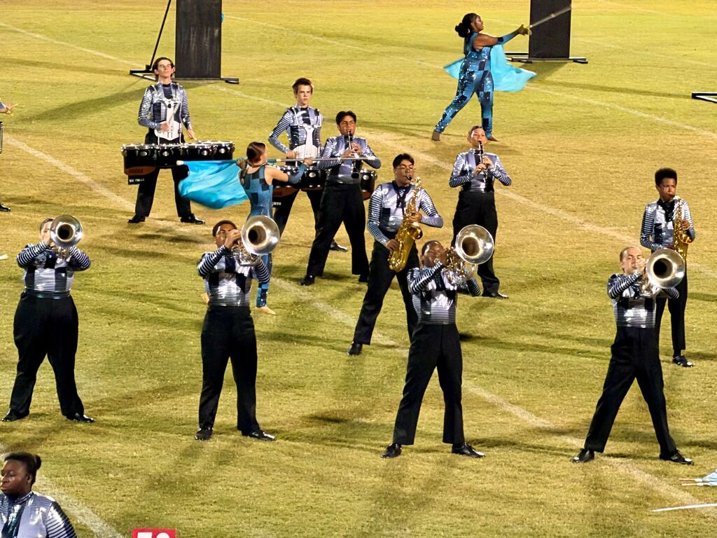 A marching band performs on a football field. Members play instruments and one dancer swings a ribbon.