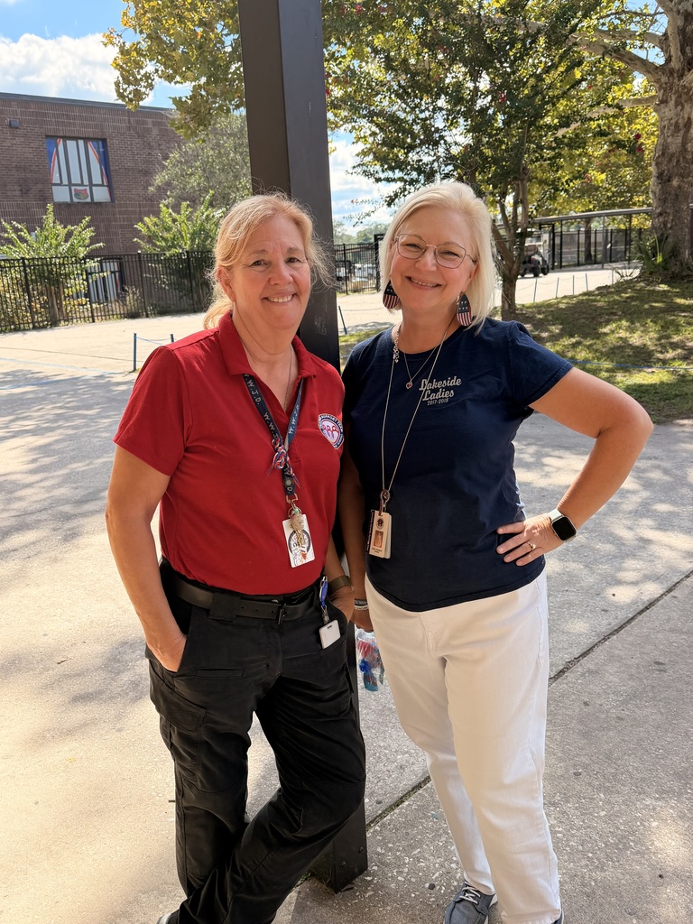 Two women stand outside in a courtyard. One wears a red shirt, the other a blue shirt. Both have eyeglasses and black belts. Trees and buildings are in the background.