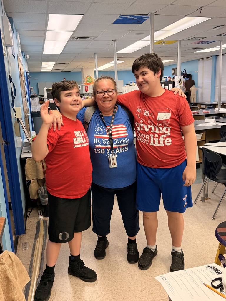 Three people in a classroom with blue walls. Two boys wear red shirts, one has a blue shirt. One boy wears black shorts, the other blue shorts.