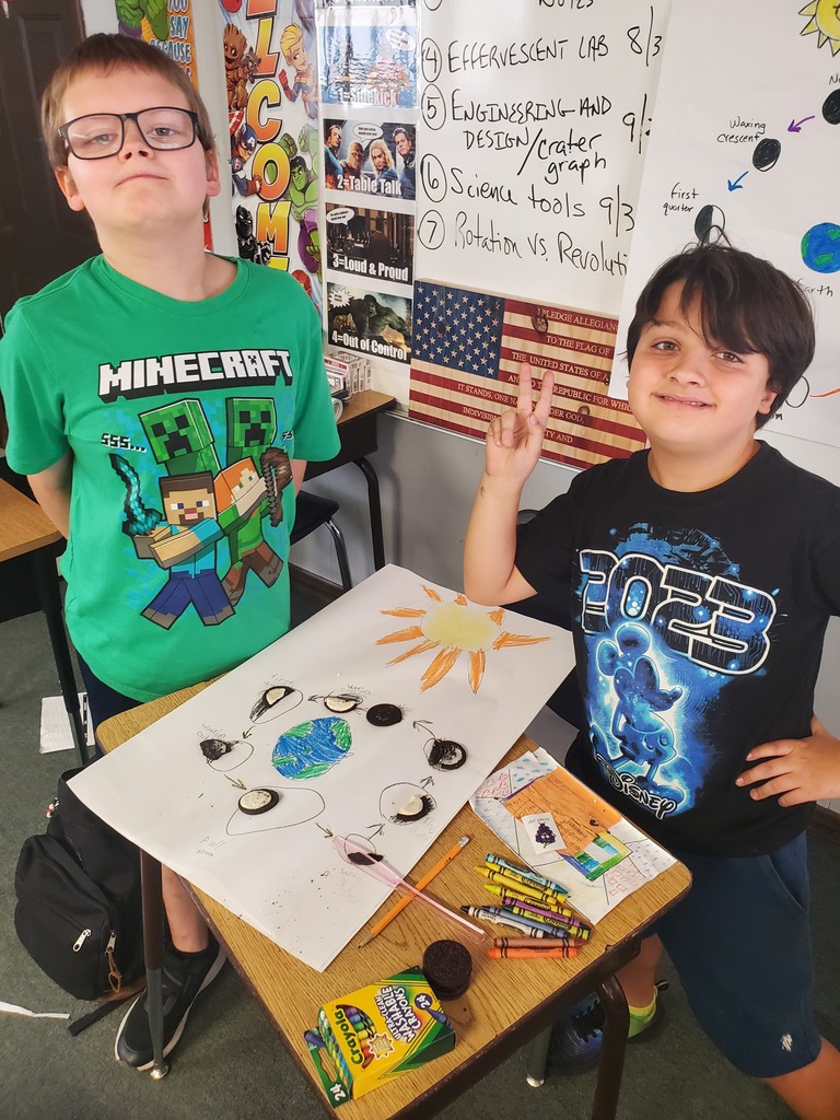 Two boys in classroom. One wears a Minecraft shirt. They stand behind a desk with art project.