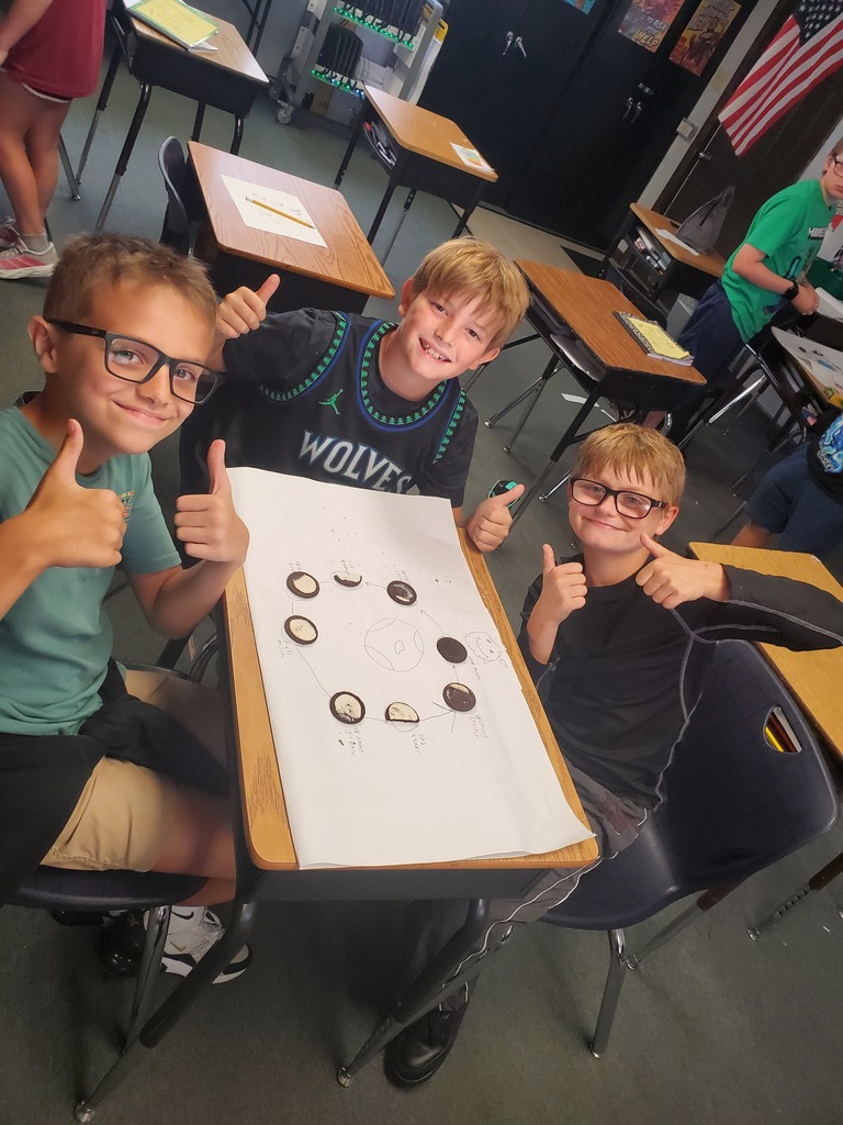 Three boys in glasses sit at a desk in a classroom, giving thumbs up. A white sheet with objects on it is on the desk.
