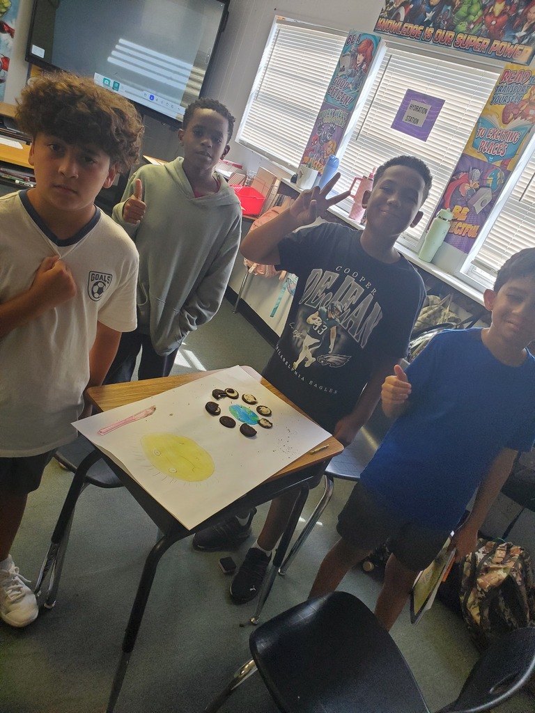 Four young students posing around a desk with a colorful drawing on paper. Bright classroom setting.