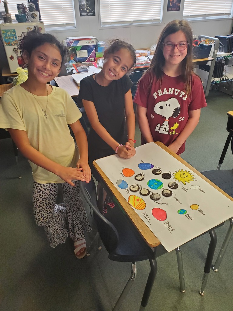 Three young girls in a classroom, one standing behind a table with a large poster displaying planets.