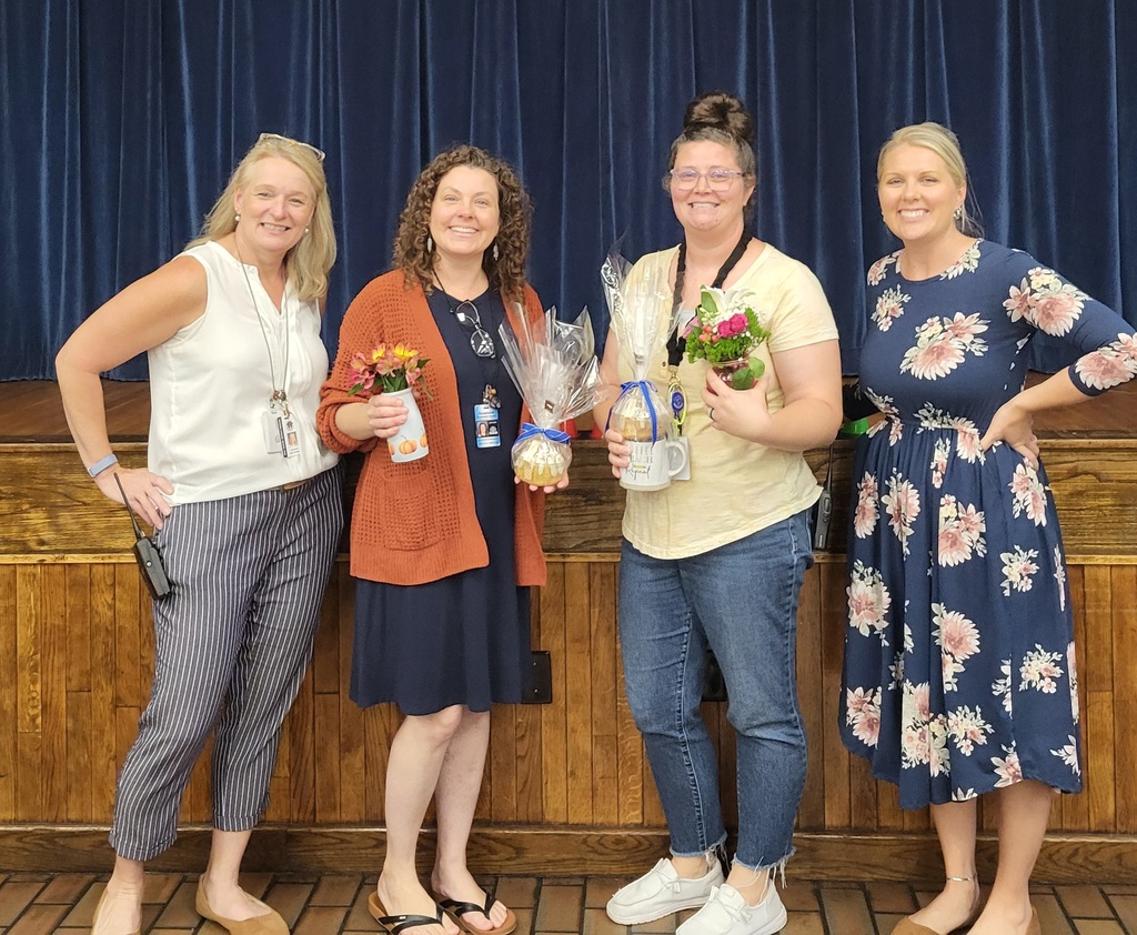 Four women, each holding a bouquet, stand in front of a wooden stage with blue curtains.